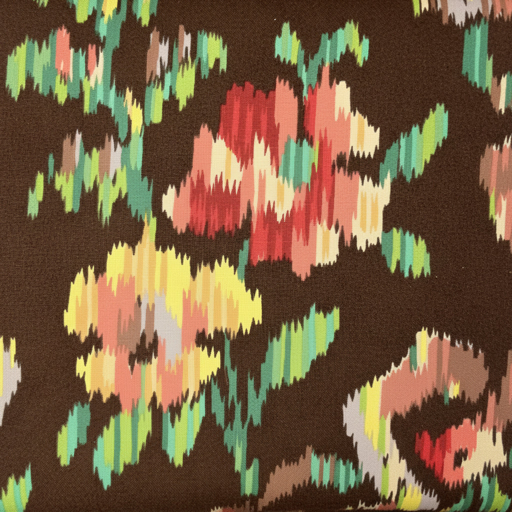 A close-up of a fabric surface with a dark charcoal base and a multicolor ikat-style floral pattern in green, yellow, pink, and beige, laid over a wooden tabletop.