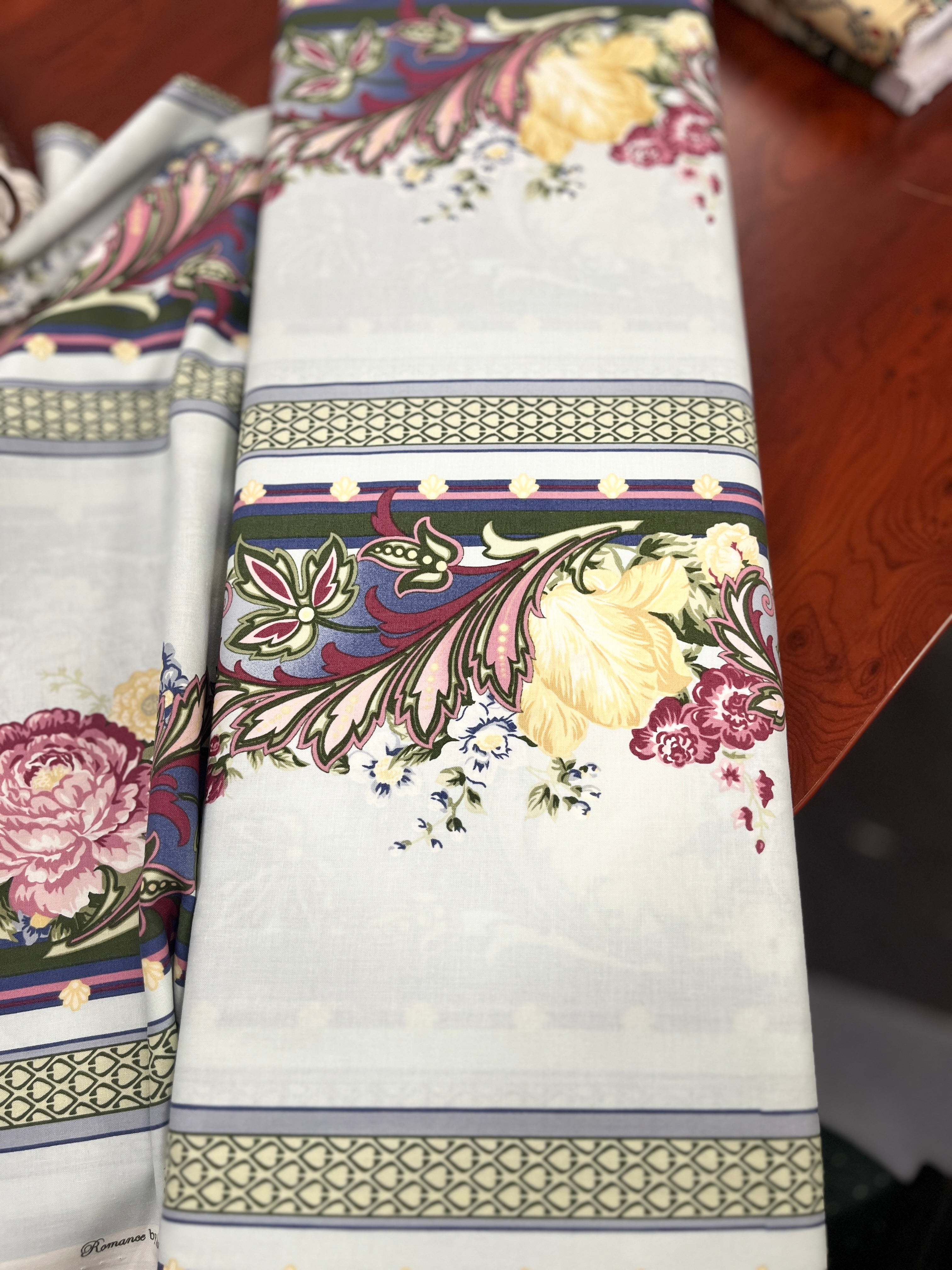 A folded white table runner with a detailed multicolor floral and botanical border pattern, displayed on a dark wooden table.