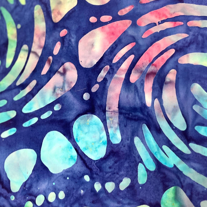 A close-up of a batik fabric featuring a swirling psychedelic pattern in shades of navy blue, mint green, light blue, and pink, with a rustic orange-brown raw edge.