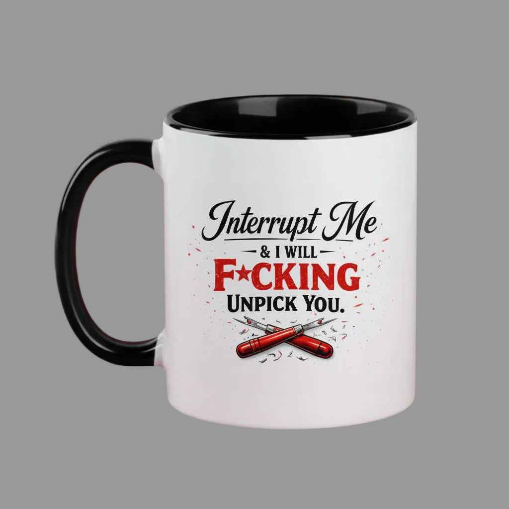 A white ceramic coffee mug with a black handle and black interior, featuring a humorous graphic that reads 'Interrupt Me & I WILL F*CKING UNPICK YOU.' in black and red lettering with crossed red tools below, set against a solid dark red background.