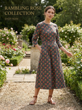 Woman in a grey Rambling Rose floral print dress with 3/4 sleeves, standing in a rose garden.