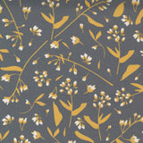 A close-up of a fabric pattern featuring delicate white flowers and mustard yellow leaves on a dark gray background, with a white ruler at the bottom showing measurement markings and the Moda brand name.