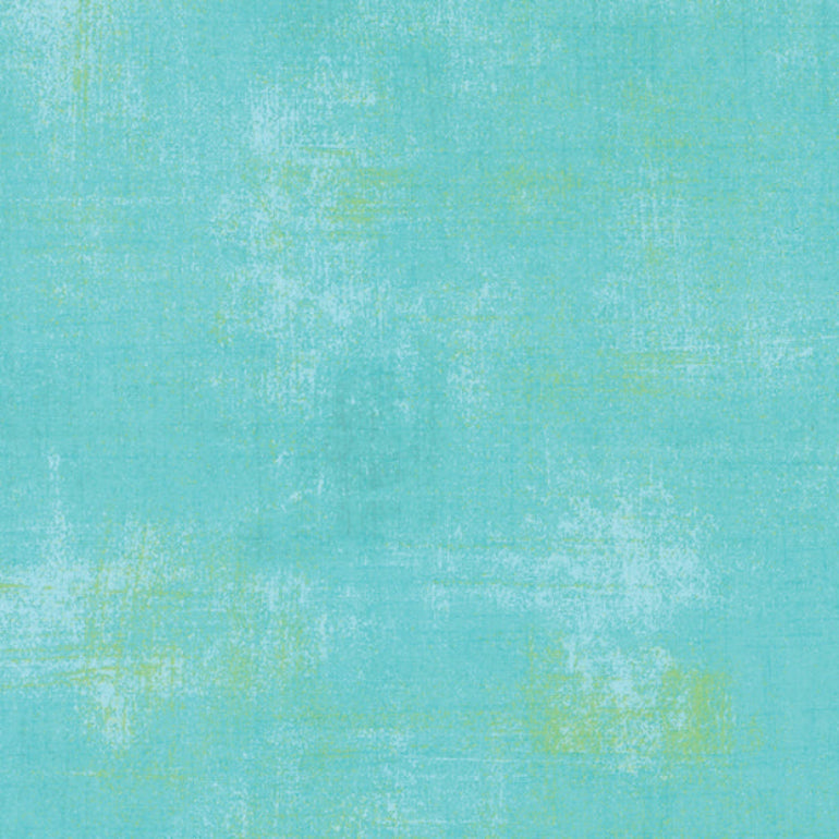 A close-up of a textured fabric swatch in turquoise with subtle pale yellow-green mottled accents, with a ruler along the bottom edge showing measurements. The brand name 'moda' and 'www.