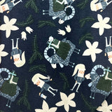 A long rectangular fabric item, likely a scarf or narrow bandana, featuring a dark navy blue background with a whimsical pattern of white flowers and small illustrated figures in green, beige, and gray, laid flat on a brown wooden surface.