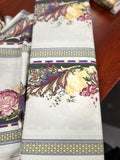 A folded white table runner with a detailed multicolor floral and botanical border pattern, displayed on a dark wooden table.