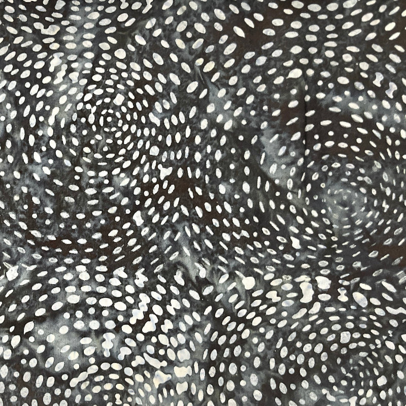 A close-up of a black fabric or material featuring a white dotted spiral pattern, with a blurred brown textured surface visible above it.