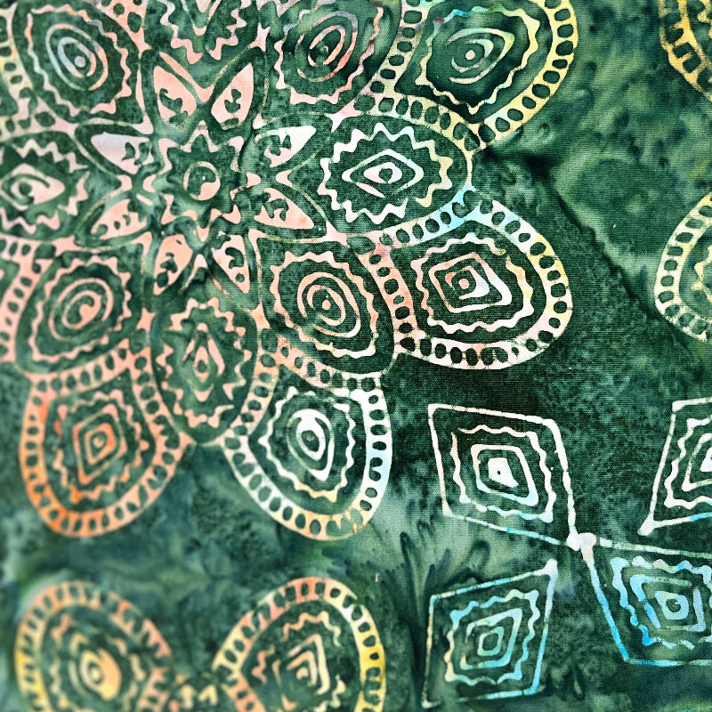 A close-up of a batik fabric featuring a green marbled background with hand-drawn mandala and geometric patterns in white, yellow, and orange.