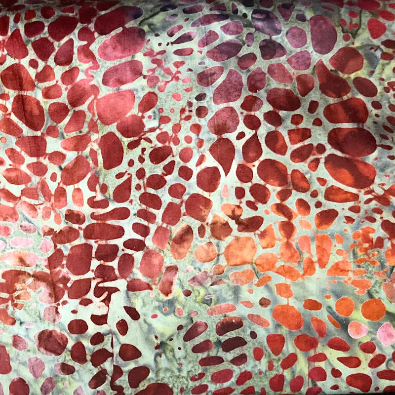 A close-up of a batik fabric with a red, orange, and beige spotted pattern, layered over other batik fabrics.