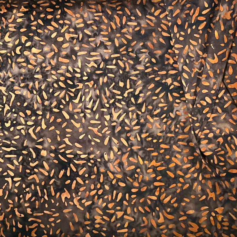 A close-up of a folded brown fabric with a dense black and orange speckled pattern, layered over other colorful patterned textiles.