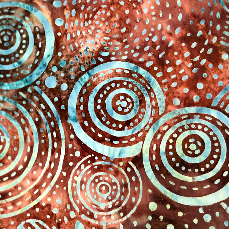A close-up of a batik fabric with a brown, orange, and blue circular dot pattern, held by a hand.