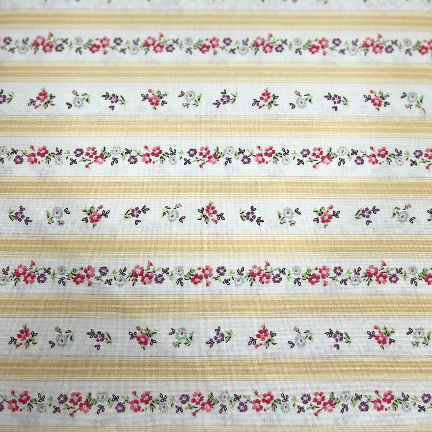 A close-up of a striped fabric or wallpaper pattern featuring horizontal bands of cream, tan, and white, decorated with repeating small floral and leaf motifs in red, green, and blue. A hand is holding the fabric at the bottom edge.