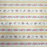 A close-up of a striped fabric or wallpaper pattern featuring horizontal bands of cream, tan, and white, decorated with repeating small floral and leaf motifs in red, green, and blue. A hand is holding the fabric at the bottom edge.