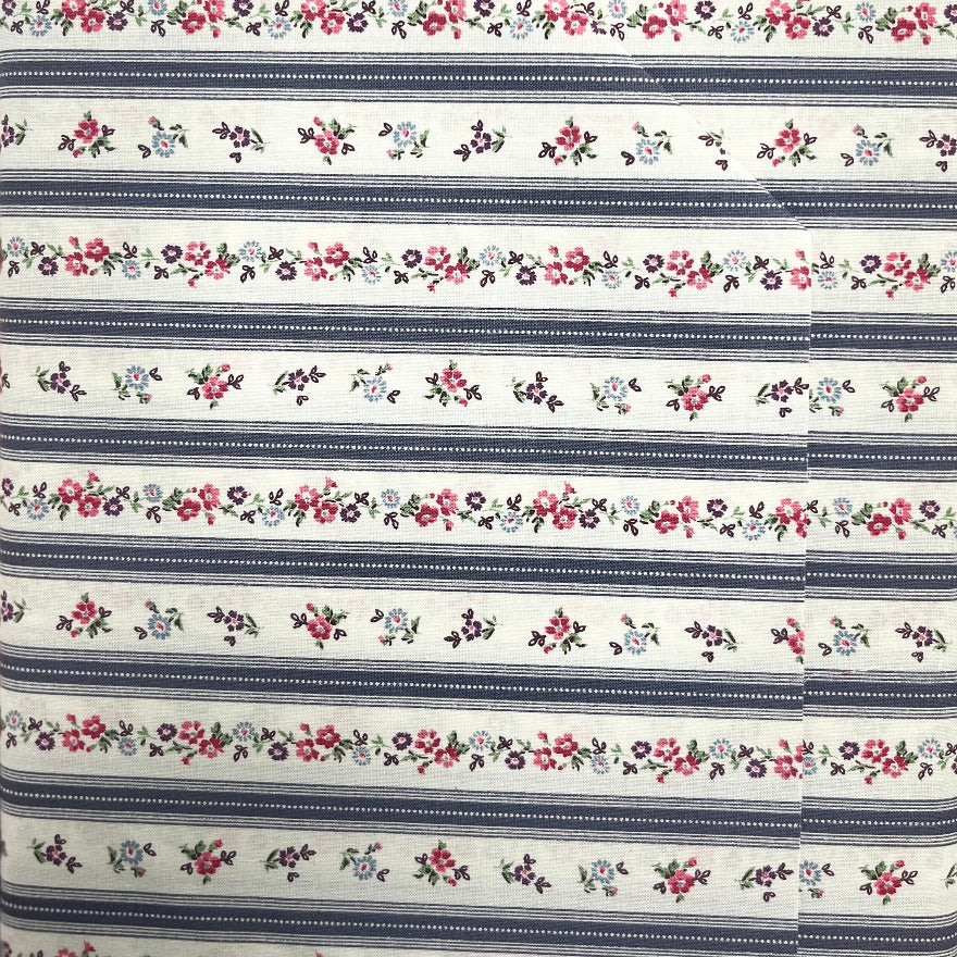 A close-up of a fabric item, likely a bed sheet or pillowcase, featuring a repeating horizontal pattern of navy blue stripes and delicate floral garlands in pink, red, and green on a white background.