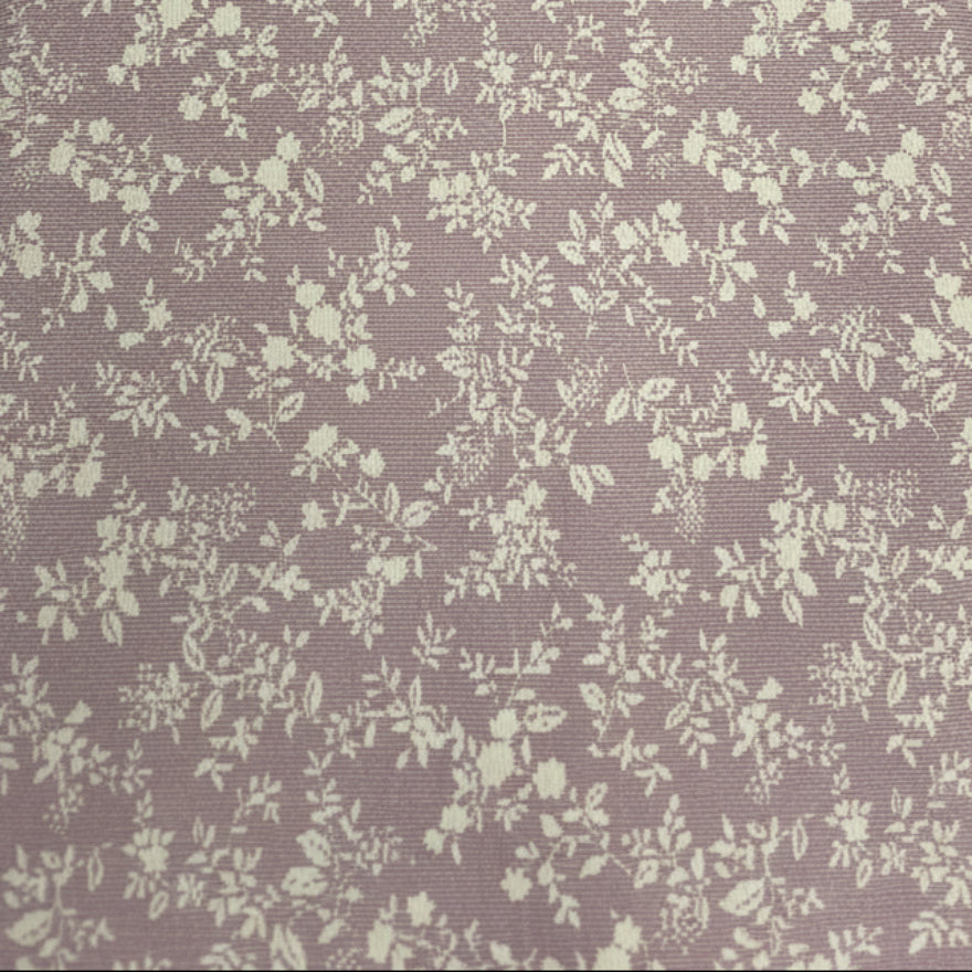 A close-up of a fabric or material sample with a small-scale floral pattern in cream on a muted dusty lavender-gray background, laid flat on a dark wooden surface.
