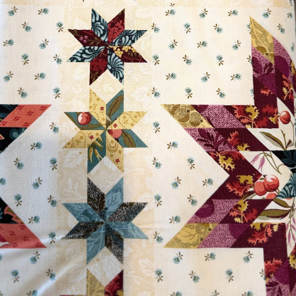 A close-up of a quilted fabric table runner featuring a vintage-style star and diamond block pattern in cream, burgundy, teal, and gold-toned fabrics.