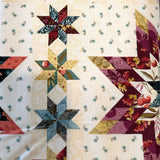 A close-up of a quilted fabric table runner featuring a vintage-style star and diamond block pattern in cream, burgundy, teal, and gold-toned fabrics.