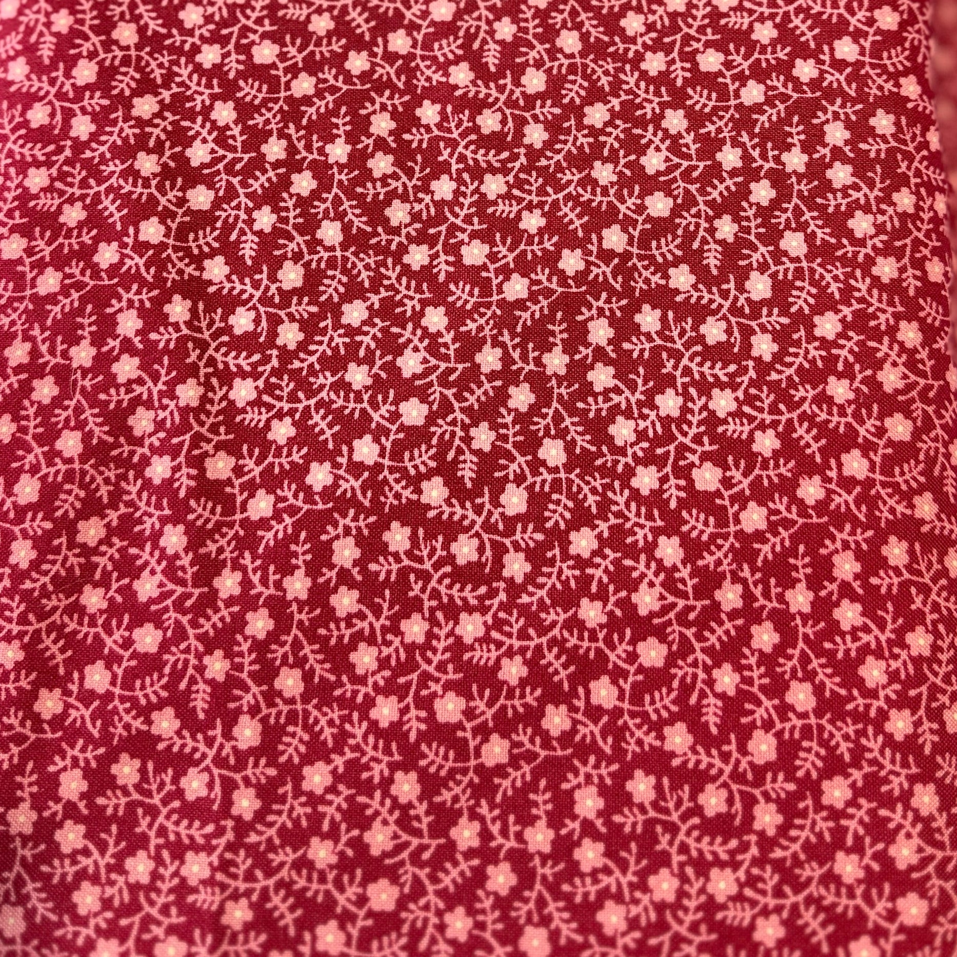 A close-up of a red fabric with a dense, small-scale white floral or ditsy print, draped vertically with a colorful, out-of-focus swatch card visible on the left edge.
