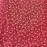 A close-up of a red fabric with a dense, small-scale white floral or ditsy print, draped vertically with a colorful, out-of-focus swatch card visible on the left edge.