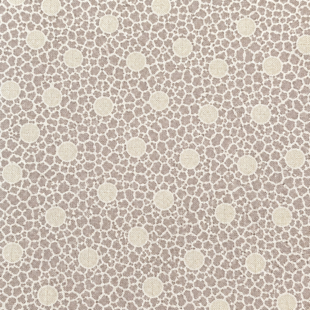 A close-up of a beige fabric or material sample with a repeating small floral pattern, held flat against a wooden surface. A purple stitched edge is visible along the right side, and another patterned fabric strip appears at the far right edge.