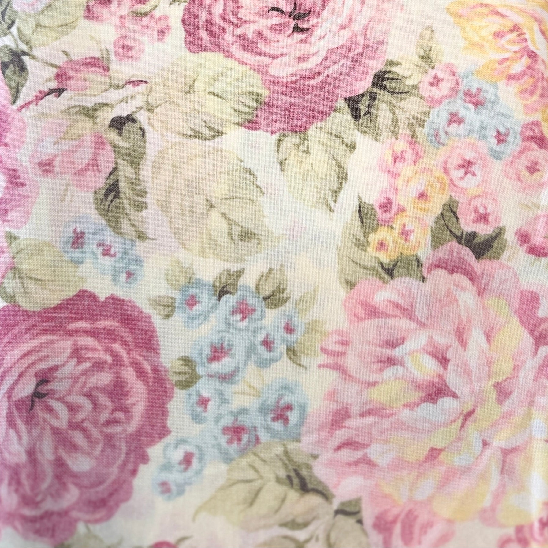 A close-up of a floral fabric with large pink and yellow roses, green leaves, and small blue flowers on a cream background.