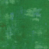 A square fabric or material swatch in mottled green tones with a subtle grid texture and a small ruler scale along the bottom edge.