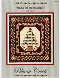 Bloom Creek 'Home for the Holidays' counted cross-stitch pattern in its original clear plastic packaging, featuring a festive quilted design with a central Christmas tree motif.