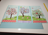 Colorful fabric panel with three whimsical trees, birds, butterflies, and flowers on a light blue background.